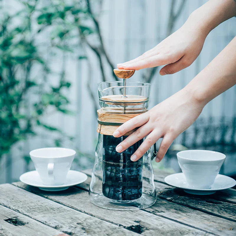 En person, der trykker ned på et trælåg af en klar glas-kaffemaskine oven på en karaffel på et træbord med to tomme hvide kopper og underkopper i nærheden. Baggrunden har grønt og et sløret hegn.