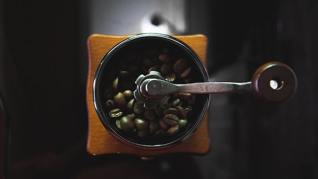 Set ovenfra og ned af en manuel kaffekværn med mørke kaffebønner indeni. Kværnen har et træhus og et metalhåndtag, placeret på en mørk overflade.