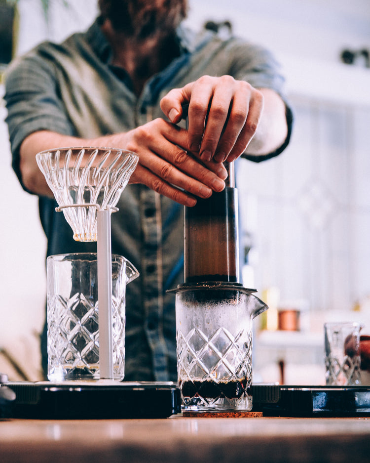 En person tilbereder kaffe ved hjælp af en Aeropress og presser enheden ned over en glasbeholder. En dryp kaffeopstilling med en glaskaraffel er i nærheden på disken. Baggrunden er lidt sløret.