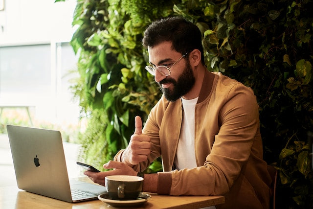 En mand med briller og skæg, iført en brun jakke, sidder ved et bord med en bærbar computer og en kaffekop. Han holder en telefon i den ene hånd og giver en tommelfinger op med den anden, omgivet af grønt.