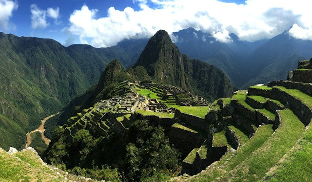 En panoramaudsigt over Machu Picchu, en gammel inkaby beliggende højt i Andesbjergene. Stedet har terrassemarker, stenstrukturer og en dramatisk top, der rejser sig i baggrunden under en delvist overskyet himmel.