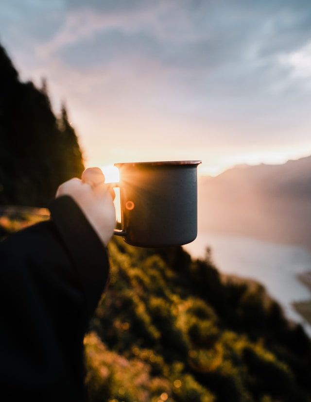 En person holder et metalkrus op mod en solopgang eller solnedgang over et naturskønt landskab med bjerge og en sø. Himlen er delvist overskyet, med sollys, der oplyser skyernes kanter og kaster et varmt skær over scenen. Forgrunden er i skygge.