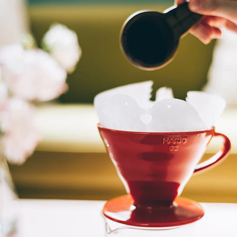 En Hario, Keramisk Drip Tragt i rød, med papirfilter, placeres på en glasbeholder. En hånd holder en sort scoop over drypperen og fremhæver dens unikke kegleform og spiralriller-design mod en blødt oplyst baggrund.