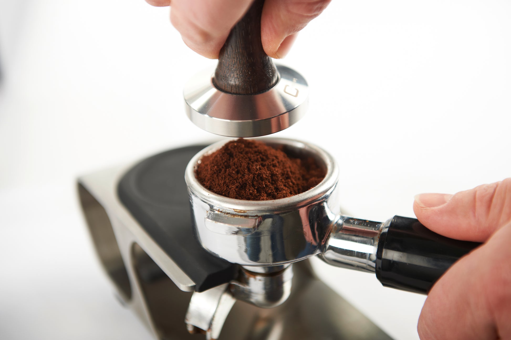 En hånd bruger en tamper til at presse kaffegrums ind i et portafilter på en espressomaskine med Joe Frex Stamper Station Pro Up, lavet af fødevaresikker silikone og rustfrit stål. Den slørede baggrund øger fokus på præcis kaffetilberedning.