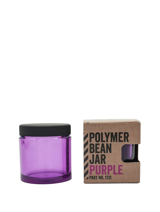 En tom, gennemsigtig lilla Comandante, Polymer Bean Jar med låg, Lilla med et sort låg er udstillet ved siden af dens brune papkasseemballage. Æsken, mærket "Comandante Polymer Bean Jar Purple Part No. 1731," har en rektangulær udskæring, der viser en del af krukken indeni, hvilket fremhæver dens fødevaresikker kvalitet.