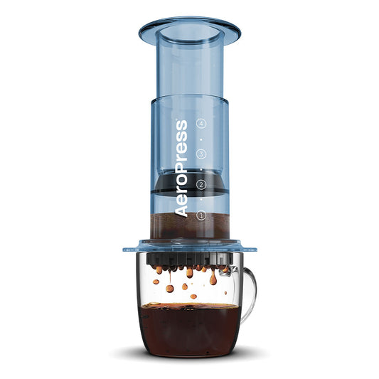 Billede af en AeroPress, Clear Limited, Kaffebrygger, Blå lavet af slidstærkt Tritan™. Den klare cylindriske anordning er vist presset ned på kaffegrums, med kaffe, der drypper ned i et klart glaskrus nedenfor. Ordet "AeroPress" er trykt lodret på enheden, hvilket fremhæver dens innovative bryggeteknologi.