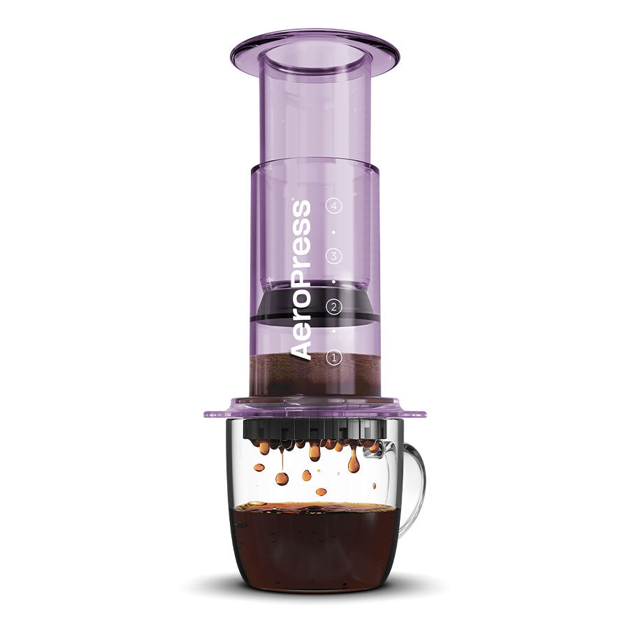 En gennemsigtig AeroPress, Clear Kaffebrygger, Lilla-limited by Aeropress er placeret over et glaskrus. Kaffe bliver brygget, hvor væske drypper fra AeroPress ned i kruset nedenfor. AeroPress, der er kendt for sin 3-i-1 bryggeteknologi, har sine komponenter samlet, inklusive stemplet og filterhætten.