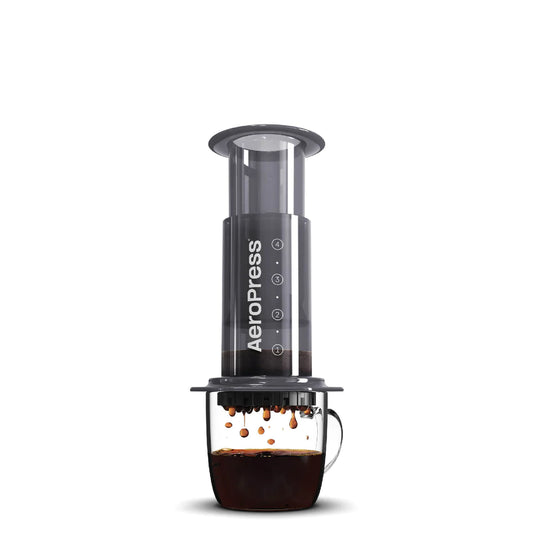 En gennemsigtig AeroPress Original kaffebrygger sidder oven på et glaskrus. Apparatet, der består af cylindre og en filterkaffebrygger, skubber friskbrygget kaffe ned i kruset nedenfor, med dråber af kaffe synlige i luften. Aeropress logoet fremhæver denne perfekte kaffeoplevelse.