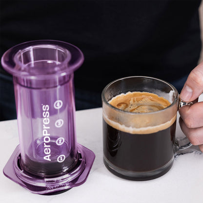 En person, der holder et glaskrus fyldt med sort kaffe ved siden af en lilla AeroPress, Clear Kaffebrygger, Lilla-limited, kendt for sin 3-i-1 bryggeteknologi, placeret på en hvid overflade og tydeligt mærket med mærkenavnet Aeropress og markeringer.