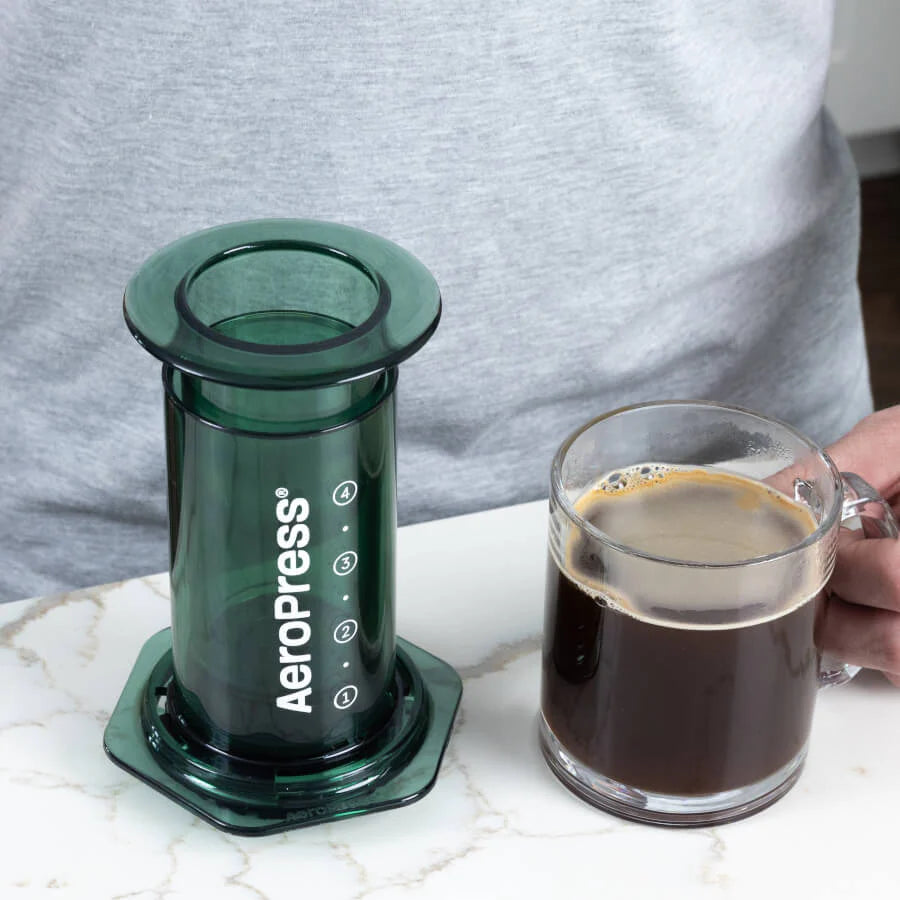 En person brygger kaffe med den grønne AeroPress Clear Limited Kaffebrygger på en marmorbordplade ved siden af et klart krus kaffe, iført en lysegrå skjorte og fremhæver Aeropress' bemærkelsesværdige bryggeteknologi.