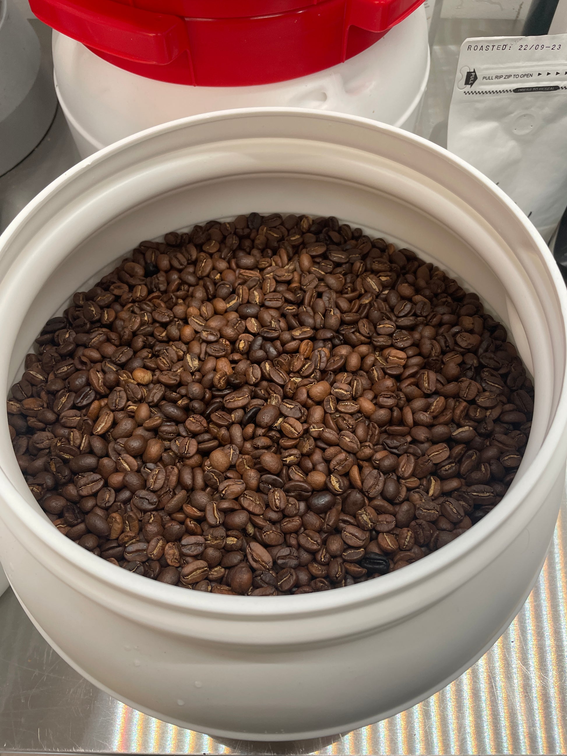 En stor hvid EverBean-beholder, nr. 05 · Honduras (b) fyldt med ristede kaffebønner sidder på en stribet overflade og byder på hints af mørk chokolade. En beholder med rødt låg står i baggrunden ved siden af en pakke mærket "Roasted.