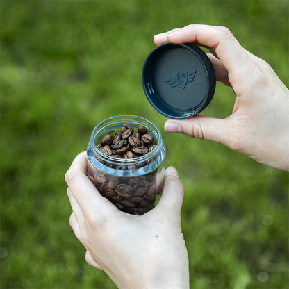 Hænder holder den åbne Comandante Polymer Bean Jar med låg, Klar stor fyldt med kaffebønner. Låget er i den ene hånd, sat mod en baggrund af frodigt grønt græs, der perfekt komplementerer friskheden og aromaen.