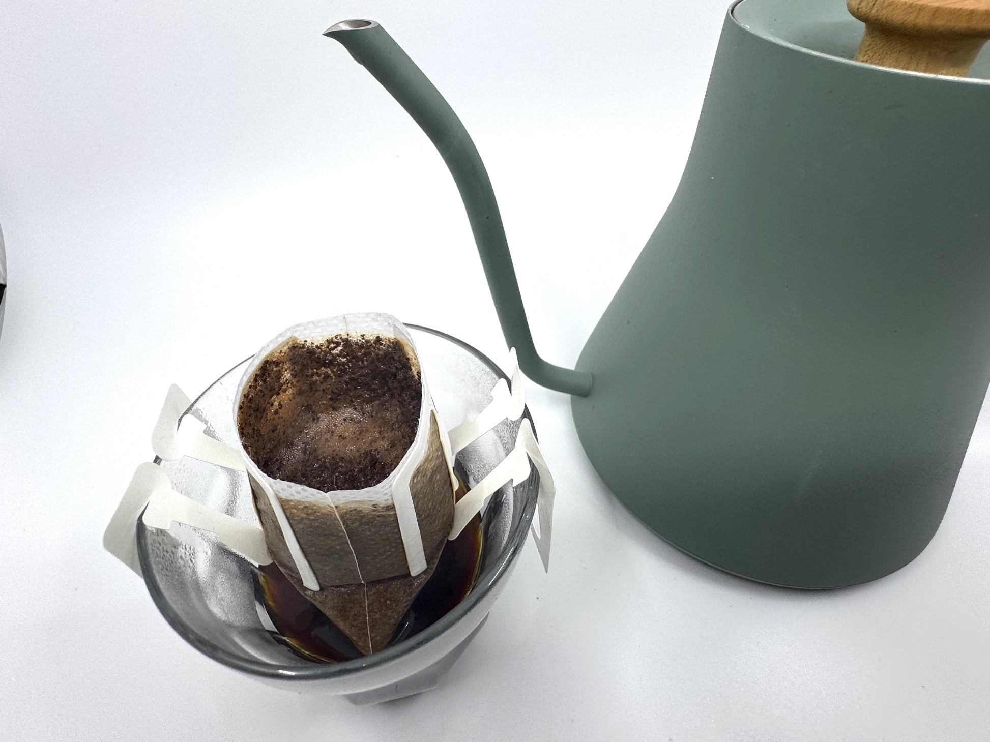 En EverBean Single Serve, nr. 02 · Brasilen sidder ved siden af en grøn kedel med et træhåndtag og brygger rig Arabica bønnen-grums i en pour-over-kaffeopstilling over en glaskaraffel. Arrangementet hviler på en hvid overflade.