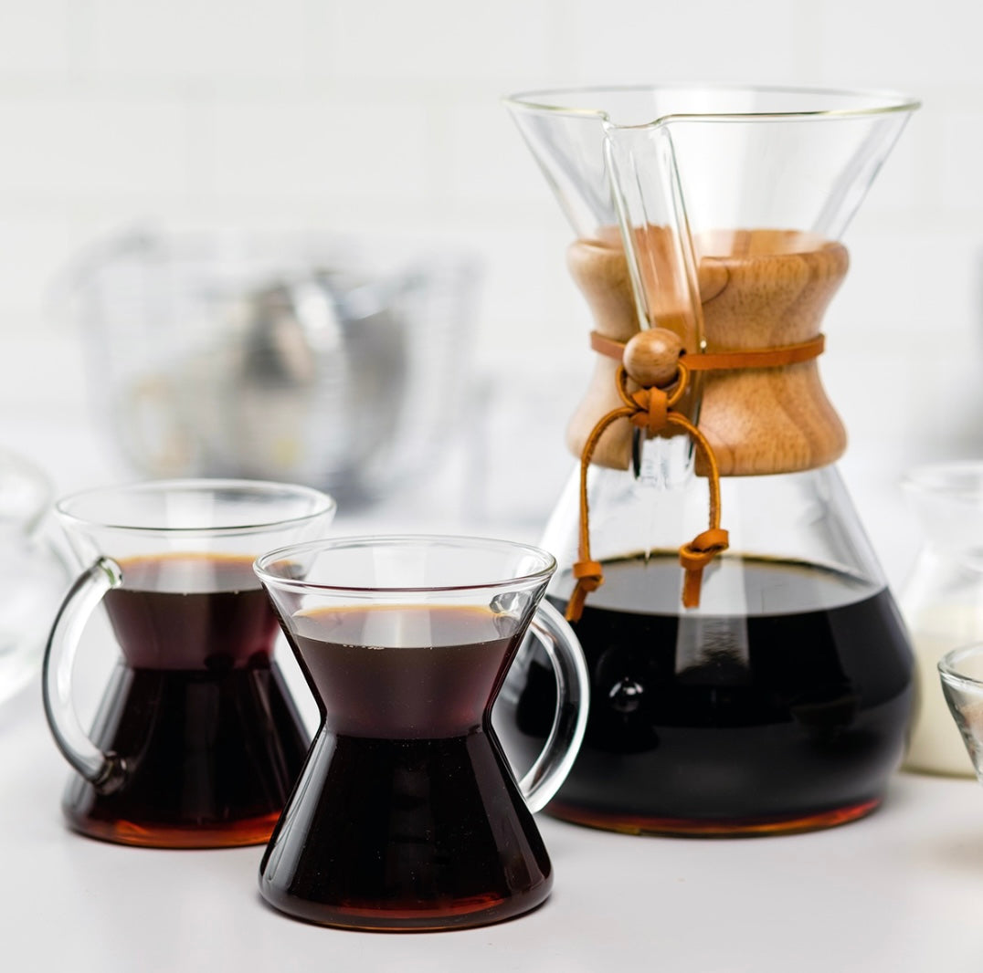 En pænt arrangeret kaffescene med en Chemex Classic Kaffebrygger 8-kopper (CM-8A) fra Chemex, ved hjælp af pour-over-teknikken, fyldt med mørk kaffe. To glaskrus, som hver indeholder kaffe, sidder ved siden af Chemex. Baggrunden inkluderer slørede køkkenting, hvilket skaber en hyggelig og indbydende stemning.