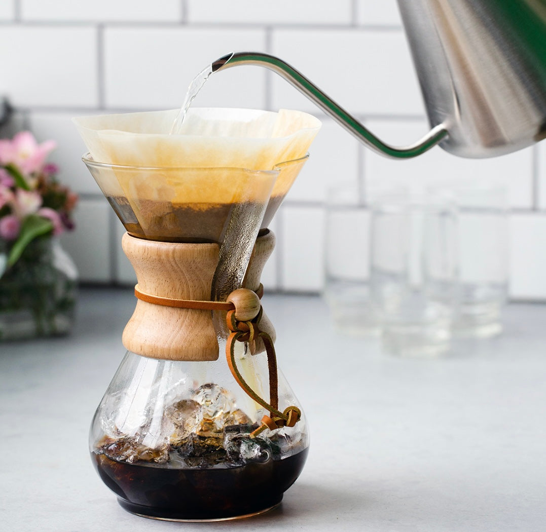 Et nærbillede af en Chemex, Classic Kaffebrygger 6-kopper (CM-6A) med en trækrave, filtreret kaffe drypper ned i glasbunden. En hånd holder en kedel i rustfrit stål og hælder varmt vand over kaffegrumset. En vase med blomster og glas er sløret i baggrunden, hvilket fremhæver elegant brygning med Chemex.