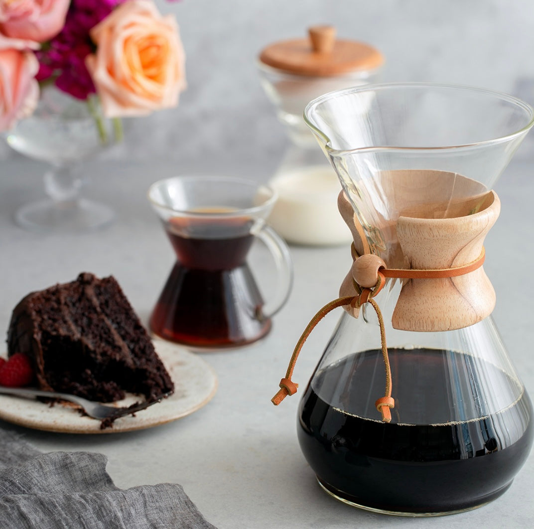 En Chemex Classic Kaffebrygger 8-kopper (CM-8A) fyldt med kaffe står på et bord ved siden af en lille karaffel kaffe, en tallerken med en skive chokoladekage og et glas mælk. I baggrunden tilføjer lyserøde og ferskenroser i en vase charmen. Chemex-modellen fremviser pour-over-teknikken perfekt.