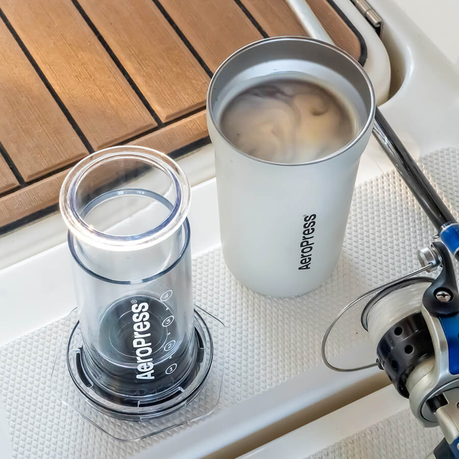 En Aeropress Go Plus kaffebrygningssetup med et krus på et båddæk. Dette Aeropress rejsekaffesystem er perfekt til dine nautiske eventyr, med en fiskestang hvilende ved siden af dem under solen.