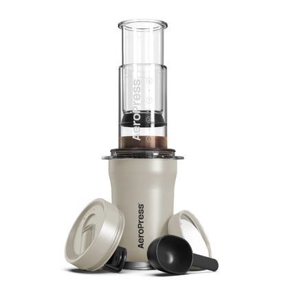 Dette rejsekaffesystem viser Aeropress, Go Plus fra Aeropress, inklusive dets pressekammer og filterhætte, sammen med en ske, omrører og rejselåg. Designet til både effektivitet og bærbarhed, er det pænt arrangeret mod en hvid baggrund.