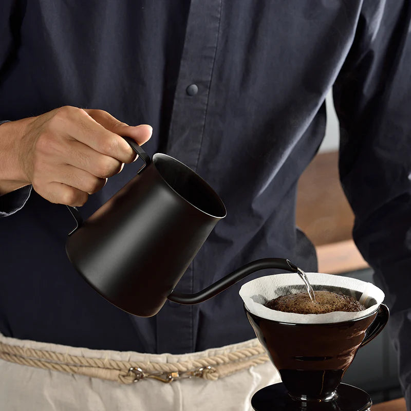 En person i mørk skjorte hælder kyndigt vand fra en Hario, Drip kedel, Kasuya Model, 500 ml i en kaffedryp med filter og jord. Drypperen, balanceret på en karaffel, tilbyder præcisionen og brugervenligheden af Tetsu Kasuya-metoden, mens de bærer et lyst forklæde.