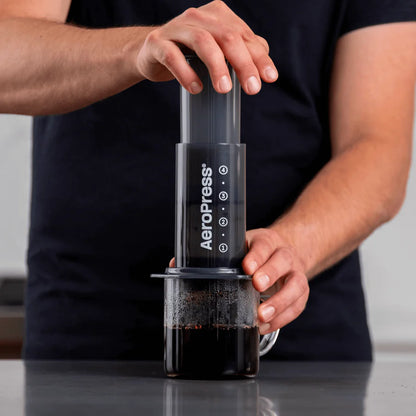 En person brygger kaffe ved hjælp af en AeroPress, Original kaffebrygger. Den ene hånd holder det øverste kammer, mens den anden trykker ned og tvinger kaffe gennem en filterkaffebrygger til et glaskrus nedenfor. Baggrunden er neutral, hvilket understreger kaffeoplevelsen og kaffeprocessen.