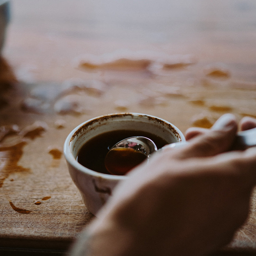 En Brewista, Professional Cupping Spoon (Titanium - Stål) holdes over en lille keramisk kop fyldt med mørk væske på en træoverflade. Lette spild og pletter er synlige rundt om koppen.