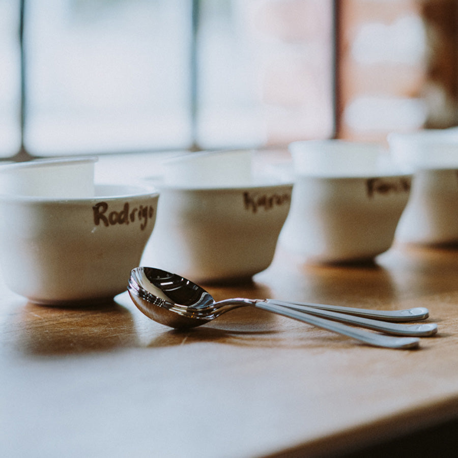 En række hvide kopper med navne skrevet i tusch sidder på et træbord, som eksemplificerer en enkel og flot stil. Foran er tre Brewista Professional Cupping Skeer (Titanium - Stål) stablet sammen. Baggrunden er blødt sløret, med et vindue synligt.