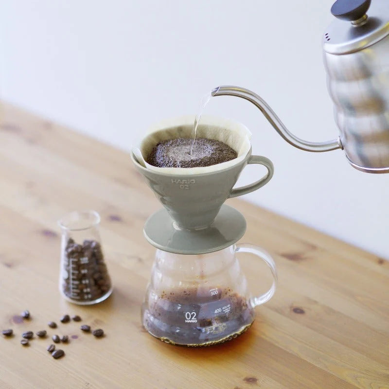 På et træbord hældes varmt vand fra en kedel i en Hario Keramisk Drip Tragt. Drypperens spiraldesign leder strømmen ind i en glaskaraffel nedenfor. I nærheden er der et bæger fyldt med kaffebønner og nogle spredt rundt.