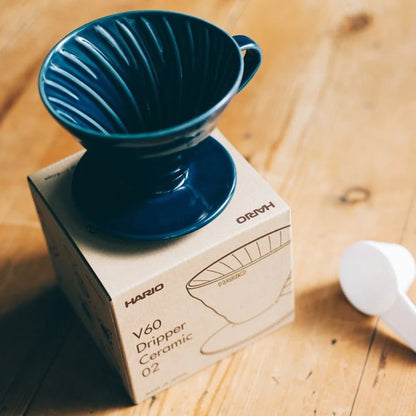 En blå keramisk Hario, Keramisk Drip Tragt kaffedrypper er placeret på en papkasse mærket "V60 Dripper Ceramic 02." Ved siden af på træbordet ligger en hvid scoop, som alle vegne kan glæde kaffeeksperter.