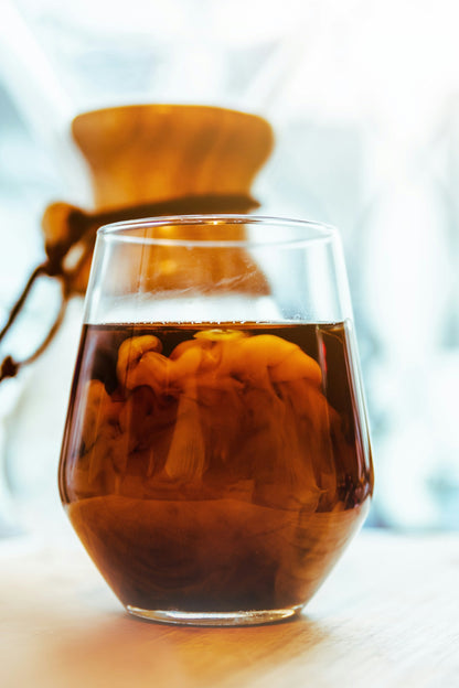 Et nærbillede af et glas fyldt med en mørk væske, muligvis EverBean ICED Coffee Blend lavet af 100% Arabica bønner, med hvirvlende creme, der skaber en marmoreret effekt. I baggrunden er en koldbryggeudstyr Chemex-stil kaffemaskine delvist synlig, lidt ude af fokus. Scenen er stærkt oplyst.