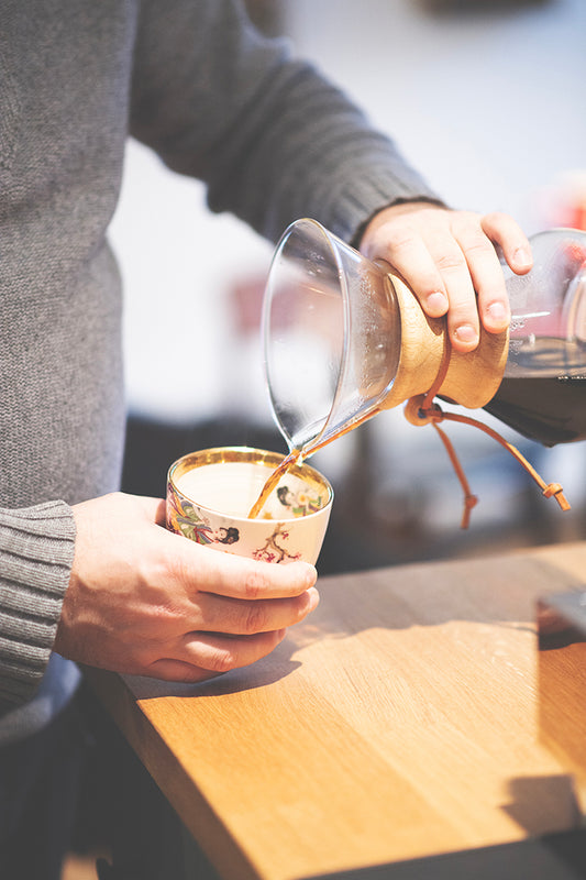 En person, der bærer en grå sweater, hælder Kenya-kaffe fra et Chemex Barista Pakke i glas (6 pax) fra Chemex & EverBean i et blomstret krus. Personens hænder og den øverste del af Chemex er synlige, og kaffen hældes på en træbordplade.